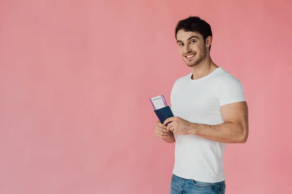 Smiling muscular man in white t-shirt holding passport and air ticket isolated on pink — Stock Photo