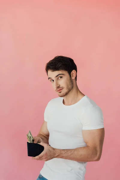 Muscular man in white t-shirt holding wallet with dollar banknotes and looking at camera isolated on pink — Stock Photo