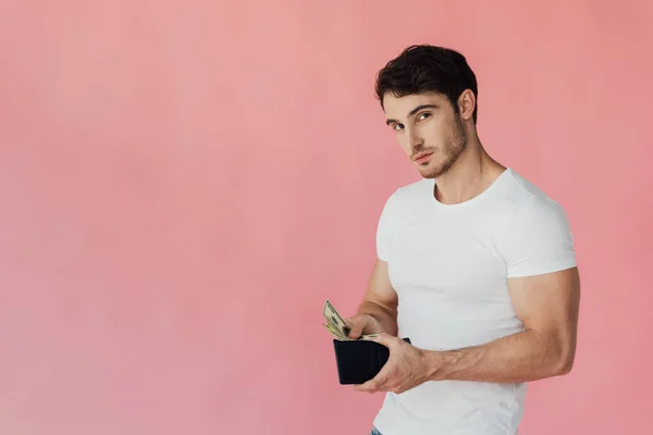 Muscular man in white t-shirt holding wallet with dollar banknotes and looking at camera isolated on pink — Stock Photo