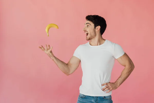 Усміхнений м'язистий чоловік у білій футболці кидає банан ізольовано на рожевому — стокове фото