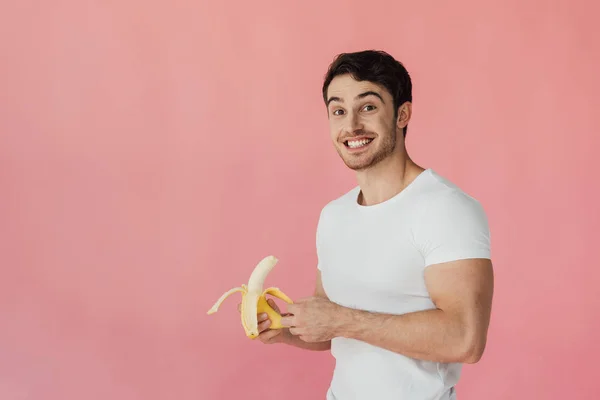 Hombre musculoso sonriente en camiseta blanca sosteniendo plátano y mirando a la cámara aislada en rosa - foto de stock