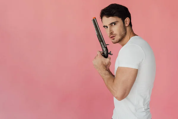 Serious muscular man in white t-shirt holding gun and looking at camera isolated on pink — Stock Photo