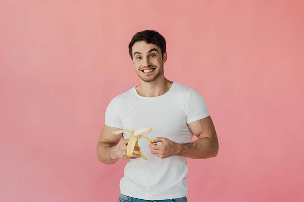 Vista frontal del hombre musculoso excitado en camiseta blanca sosteniendo plátano aislado en rosa - foto de stock