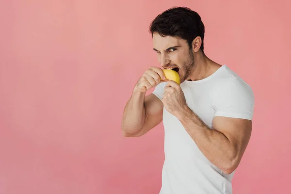 Голодный мускулистый мужчина в белой футболке ест банан изолированный на розовый — стоковое фото