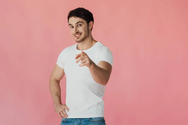 Smiling muscular man in white t-shirt standing with hand on hip and pointing with finger at camera isolated on pink — Stock Photo