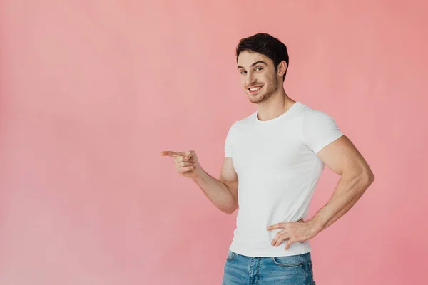 Uomo muscoloso sorridente in t-shirt bianca in piedi con mano sull'anca e punta con dito isolato su rosa — Foto stock