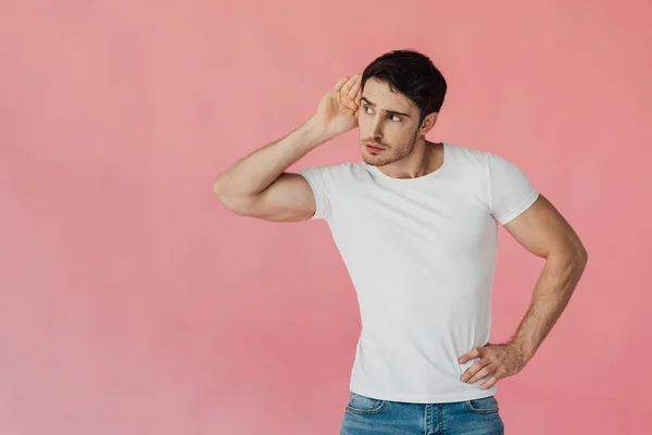 Hombre musculoso en camiseta blanca sosteniendo la palma cerca de la oreja y escuchando aislado en rosa - foto de stock