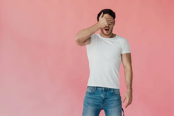 Vorderansicht eines schockierten muskulösen Mannes in weißem T-Shirt, der die Augen mit einer Hand isoliert auf rosa verdeckt — Stockfoto