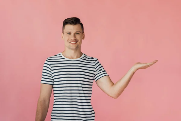 Vorderseite eines lächelnden Mannes in gestreiftem T-Shirt, der mit einer Hand isoliert auf pink zeigt — Stockfoto
