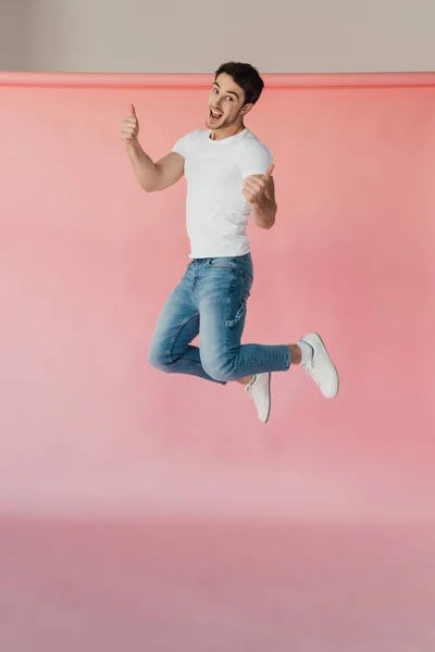 Homem muscular sorridente em t-shirt branca e jeans pulando e mostrando polegares para cima em rosa — Fotografia de Stock
