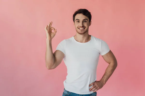 Vista frontale dell'uomo muscoloso sorridente in t-shirt bianca che mostra un segno positivo isolato sul rosa — Foto stock