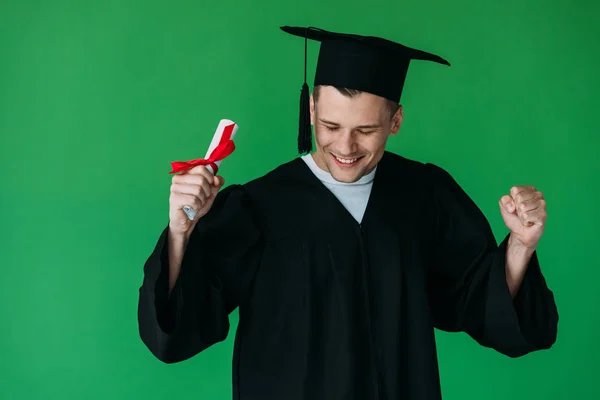 Estudiante sonriente en gorra académica sosteniendo diploma con cinta roja y mostrando sí gesto aislado en verde - foto de stock