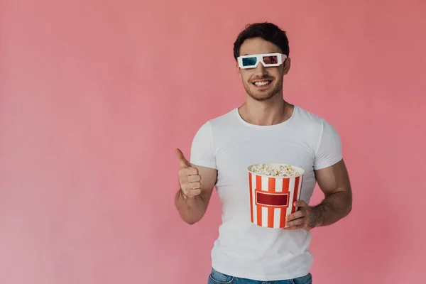 Вид спереду усміхненого м'язового чоловіка в 3d окулярах, що тримає попкорн і показує великий палець вгору ізольовано на рожевому — стокове фото