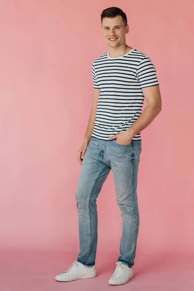 Ganzkörperansicht eines lächelnden Mannes in Jeans und gestreiftem T-Shirt mit der Hand in der Tasche, der in die Kamera auf rosa schaut — Stockfoto