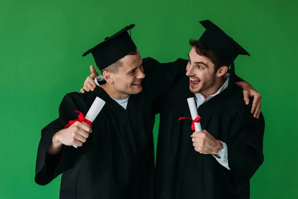 Два счастливых студента в академических шапках, имеющих дипломы и обнимающих изолированных на зеленый — стоковое фото