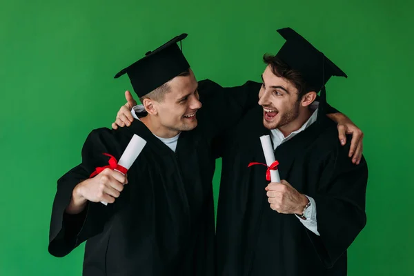 Deux étudiants heureux dans des casquettes académiques détenant des diplômes et embrassant isolé sur vert — Photo de stock