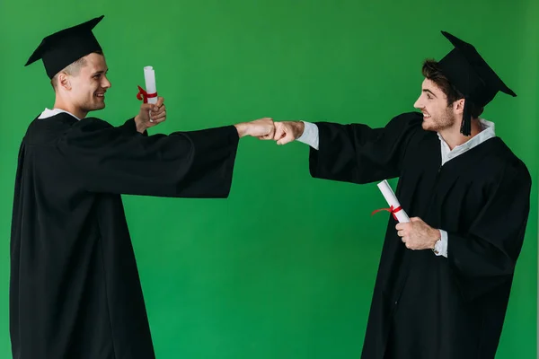 Due studenti felici in berretti accademici in possesso di diplomi e pugni toccanti isolati su verde — Foto stock