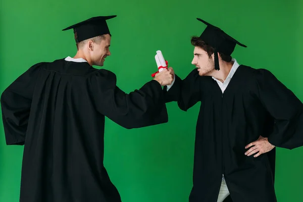 Two students in academic caps holding diplomas and looking at each other isolated on green — Stock Photo