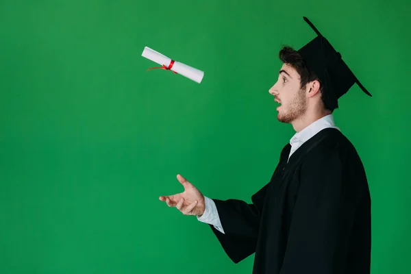 Side view of surprised student in academic cap throwing up diploma with red ribbon isolated on green — Stock Photo