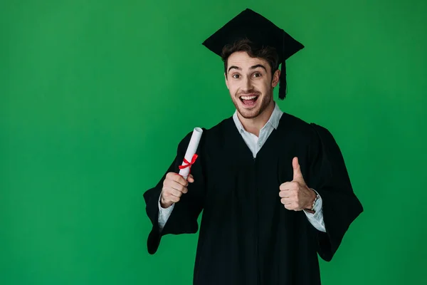Excited student in academic cap holding diploma with red ribbon and showing thumb up isolated on green — Stock Photo