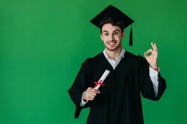 Вид спереду студента в академічній шапці, що тримає диплом з червоною стрічкою і показує добре знак ізольовано на зеленому — стокове фото