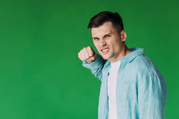 Сердитий молодий чоловік у сорочці тримає кулак і дивиться на камеру ізольовано на зеленому — стокове фото