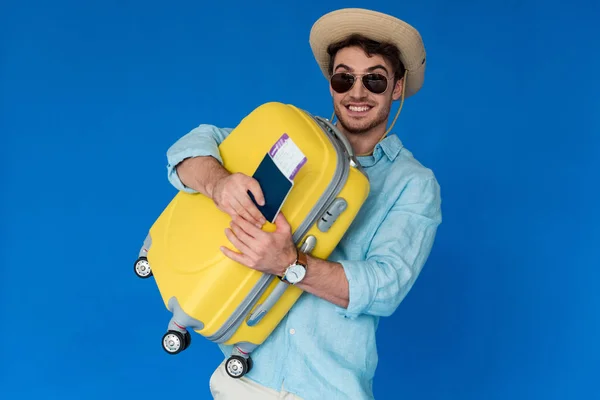 Viajero emocionado en sombrero de safari y gafas de sol con maleta amarilla y pasaporte con billete de avión aislado en azul - foto de stock