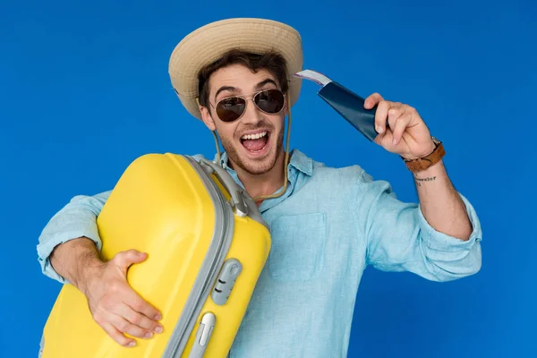Viajero emocionado en sombrero de safari y gafas de sol con maleta amarilla y pasaporte con billete de avión aislado en azul - foto de stock