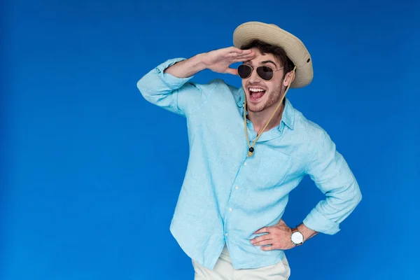Excited tourist in safari hat and sunglasses laughing and looking in distance isolated on blue — Stock Photo