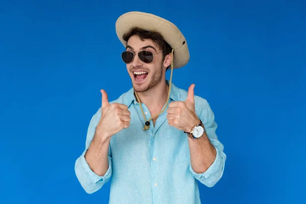 Viajero emocionado en sombrero de safari y gafas de sol que muestran los pulgares hacia arriba aislado en azul - foto de stock