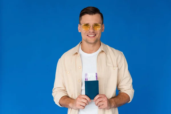 Vista frontal del viajero sonriente en gafas de sol con pasaporte con billete de avión aislado en azul - foto de stock