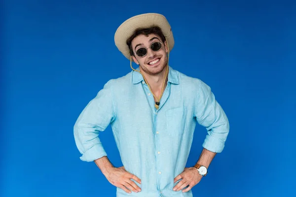 Front view of smiling traveler in safari hat and sunglasses standing with hands on hips isolated on blue — Stock Photo