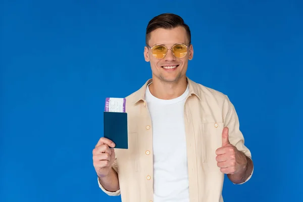 Vista frontal del turista sonriente en gafas de sol con pasaporte y mostrando el pulgar hacia arriba con billete de avión aislado en azul - foto de stock
