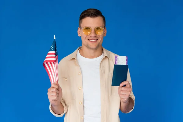Вид улыбающегося туриста в солнечных очках, держащего паспорт с авиабилетом и американским флагом, изолированным на голубом — стоковое фото