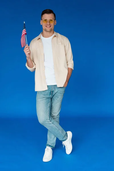 Full length view of smiling man in sunglasses standing with hand in pocket and holding american flag on blue — Stock Photo