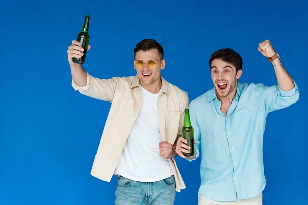 Dos amigos excitados sosteniendo botellas de cerveza y mostrando sí gesto aislado en azul - foto de stock