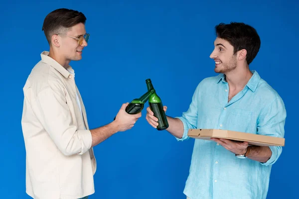 Dos amigos sonrientes tintinean botellas de cerveza y se miran aislados en azul - foto de stock