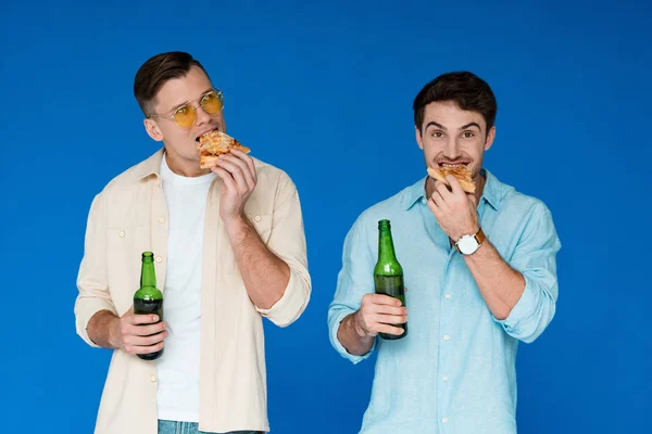 Vista frontale di due amici che tengono bottiglie di birra e mangiano pizza isolata sul blu — Foto stock