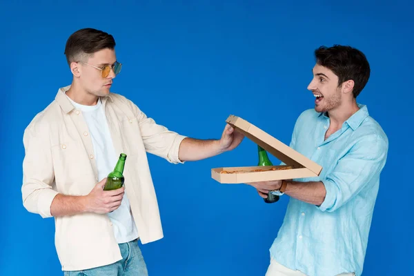 Two friends holding bottles of beer and pizza isolated on blue — Stock Photo