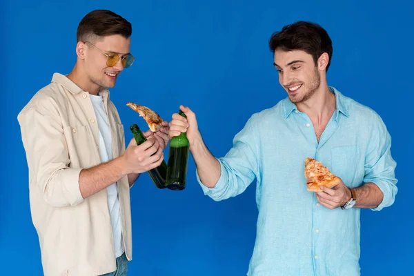 Two smiling friends holding bottles of beer and slices of pizza isolated on blue — Stock Photo