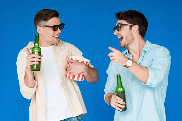 Two smiling friends in 3d glasses holding bottles of beer and popcorn isolated on blue — Stock Photo
