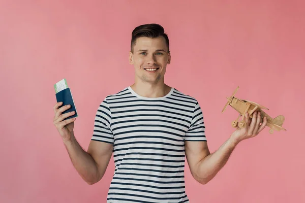 Vista frontal del sonriente turista sosteniendo pasaporte con billete de avión y avión de juguete de madera aislado en rosa - foto de stock