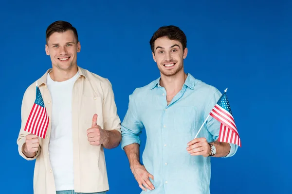 Vista frontal de dois amigos sorridentes segurando bandeiras americanas e mostrando polegar para cima isolado em azul — Fotografia de Stock
