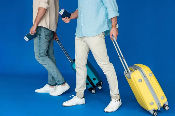 Cropped view of two tourists holding suitcases and passports with air tickets on blue — Stock Photo