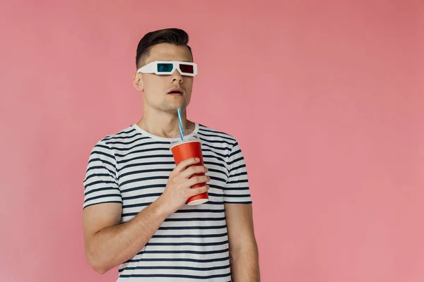 Surprised young man in 3d glasses and striped t-shirt holding beverage and looking away isolated on pink — Stock Photo
