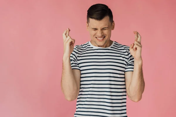 Front view of worried young man in striped t-shirt with crossed fingers isolated on pink — Stock Photo