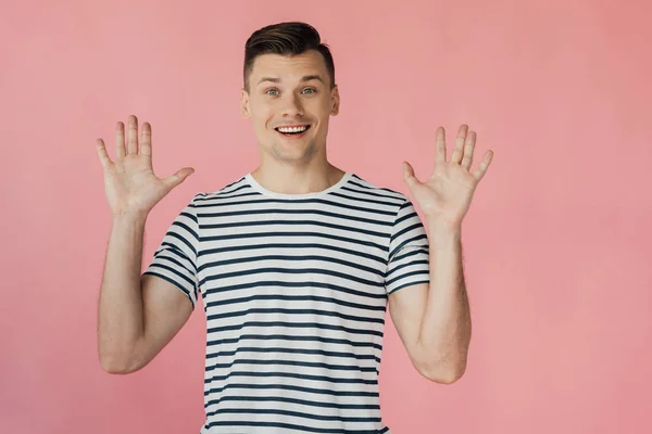 Vue de face du jeune homme souriant excité en t-shirt rayé debout avec les mains en l'air isolé sur rose — Photo de stock