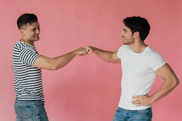 Deux amis souriants en t-shirts se touchant les poings et se regardant isolés sur rose — Photo de stock