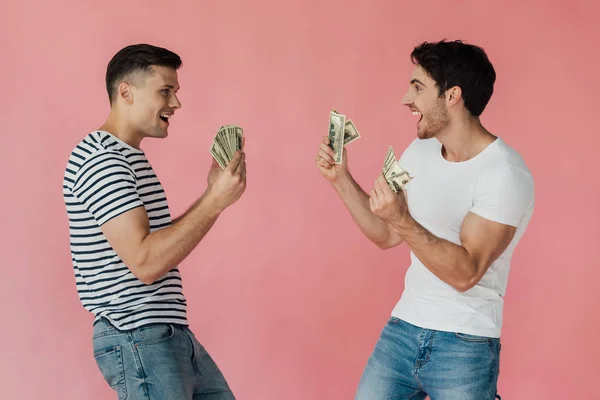 Two excited friends with dollar banknotes looking at each other isolated on pink — Stock Photo