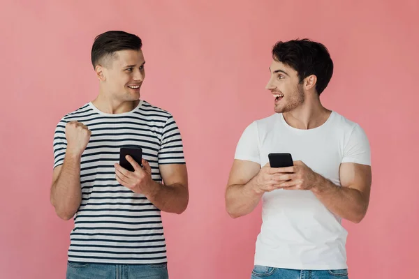 Dos hombres sonrientes usando teléfonos inteligentes y mirándose aislados en rosa - foto de stock